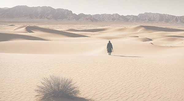 A lone person walking across vast sand dunes in a desert with mountains in the background.